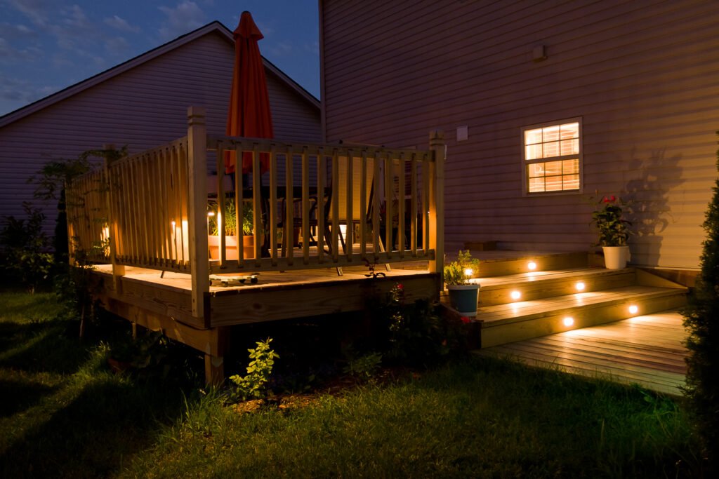 Cozy backyard deck at night, illuminated by soft step lights and warm glow from lanterns, surrounded by plants