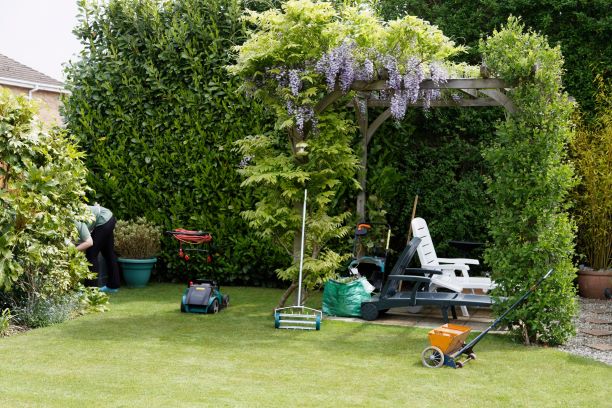 Garden with lawn aeration tools, a pergola covered in flowering vines, and gardening equipment, surrounded by lush greenery