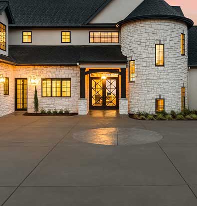 Elegant hardscape featuring a smooth concrete driveway leading to a stone-clad home with a turret and warm exterior lighting