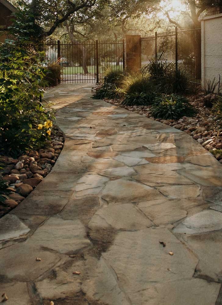 Flagstone pathway bordered by natural stone and lush greenery, leading to a gated entrance in a peaceful outdoor setting