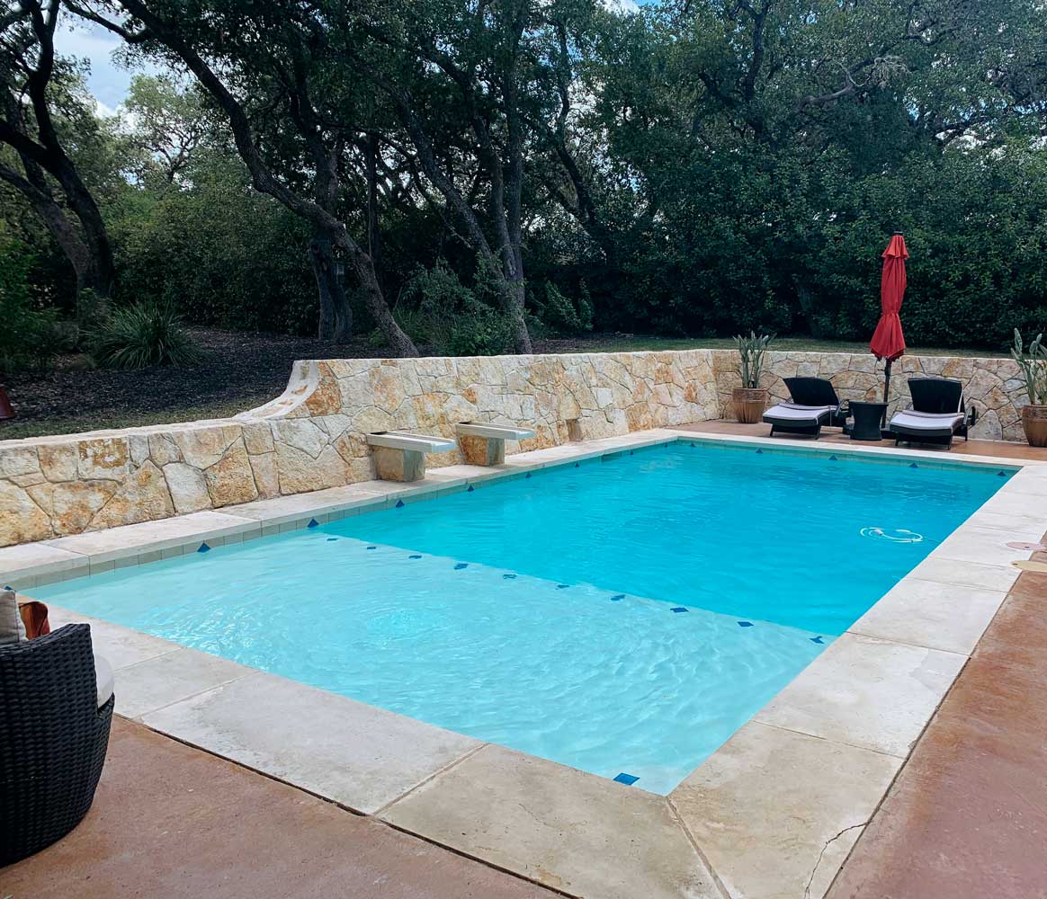 Serene pool area featuring stone hardscapes, lounge chairs, and lush trees in the background, with a red umbrella for shade