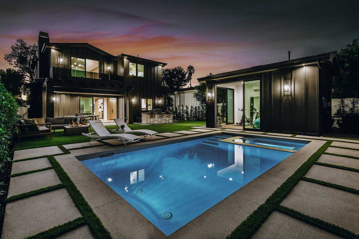 Modern backyard at night featuring illuminated hardscapes, a glowing pool, lounge chairs, and a sleek dark-toned house