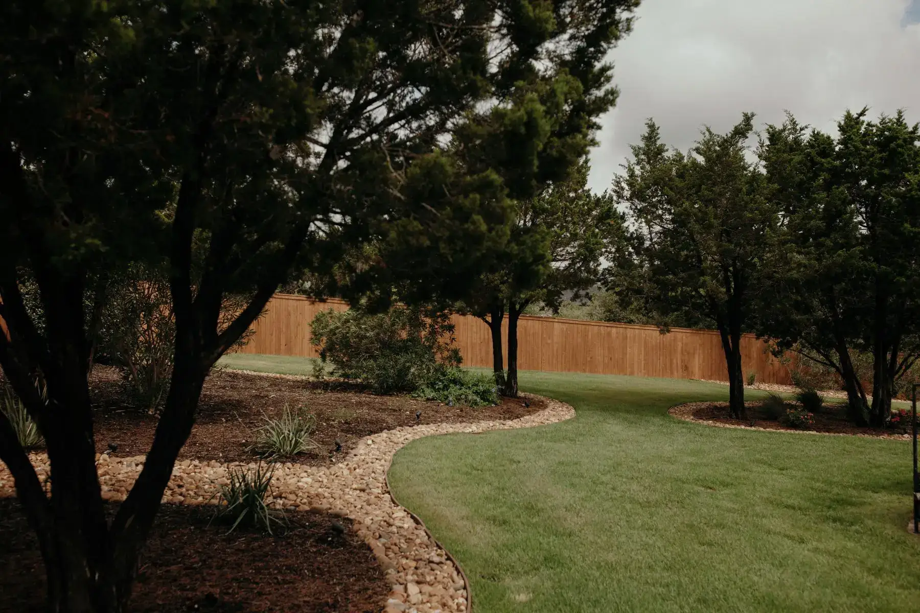 Backyard with mature trees, a well-manicured lawn, and stone-edged garden beds, emphasizing natural Softscapes