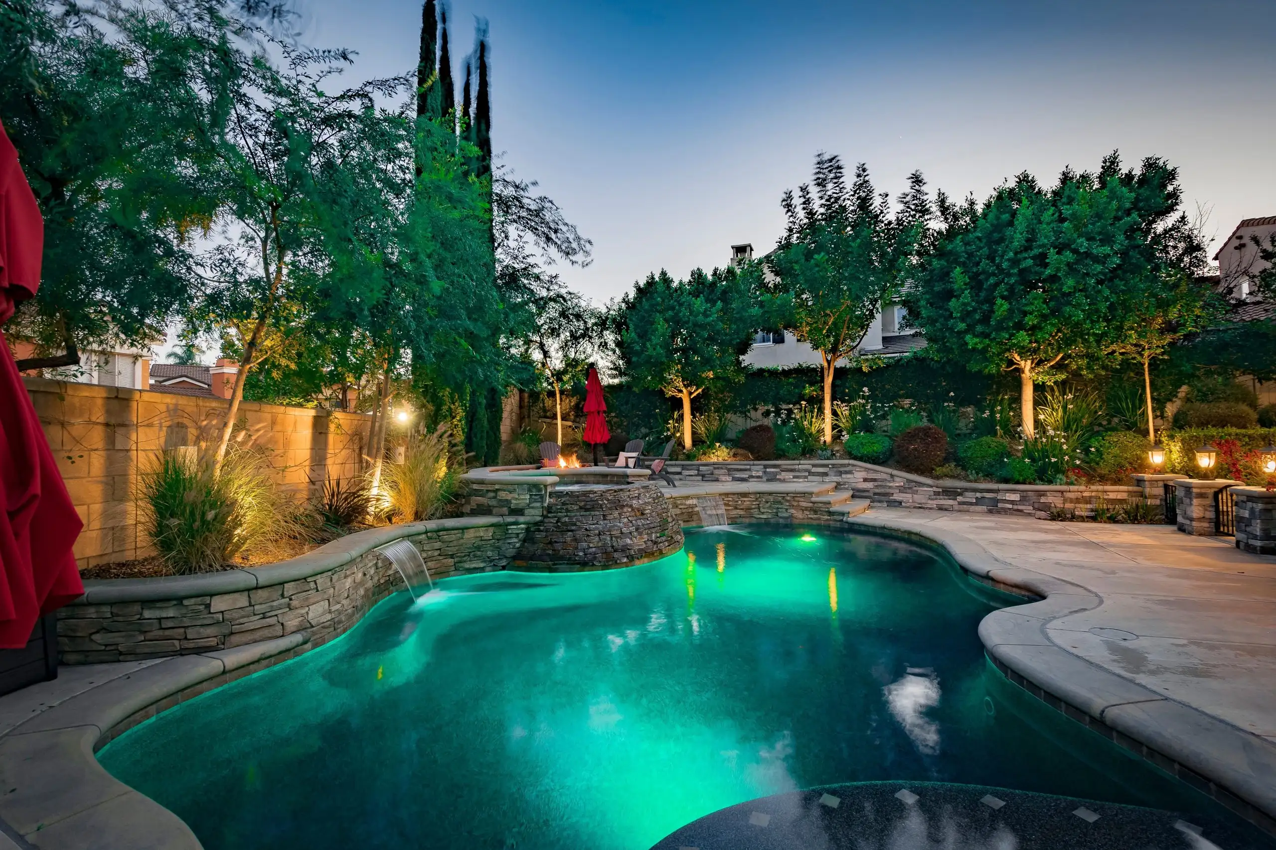 Beautifully lit backyard pool area with stone retaining walls and lush landscaping, showcasing elegant Hardscapes