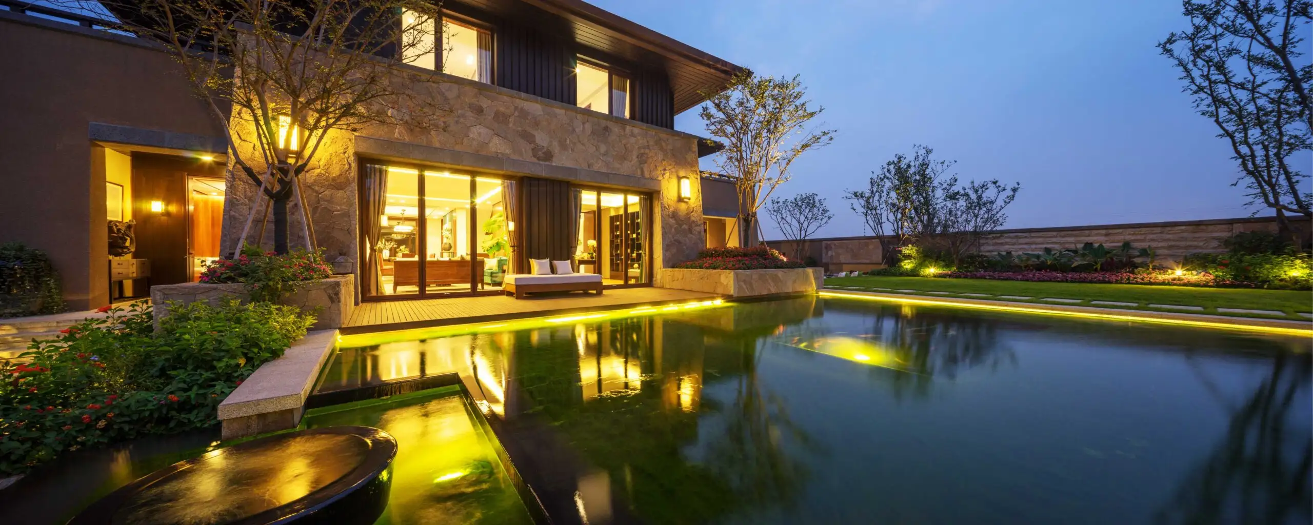 Luxurious backyard view at dusk featuring a reflective pond, softscapes with lush plants, and a well-lit patio