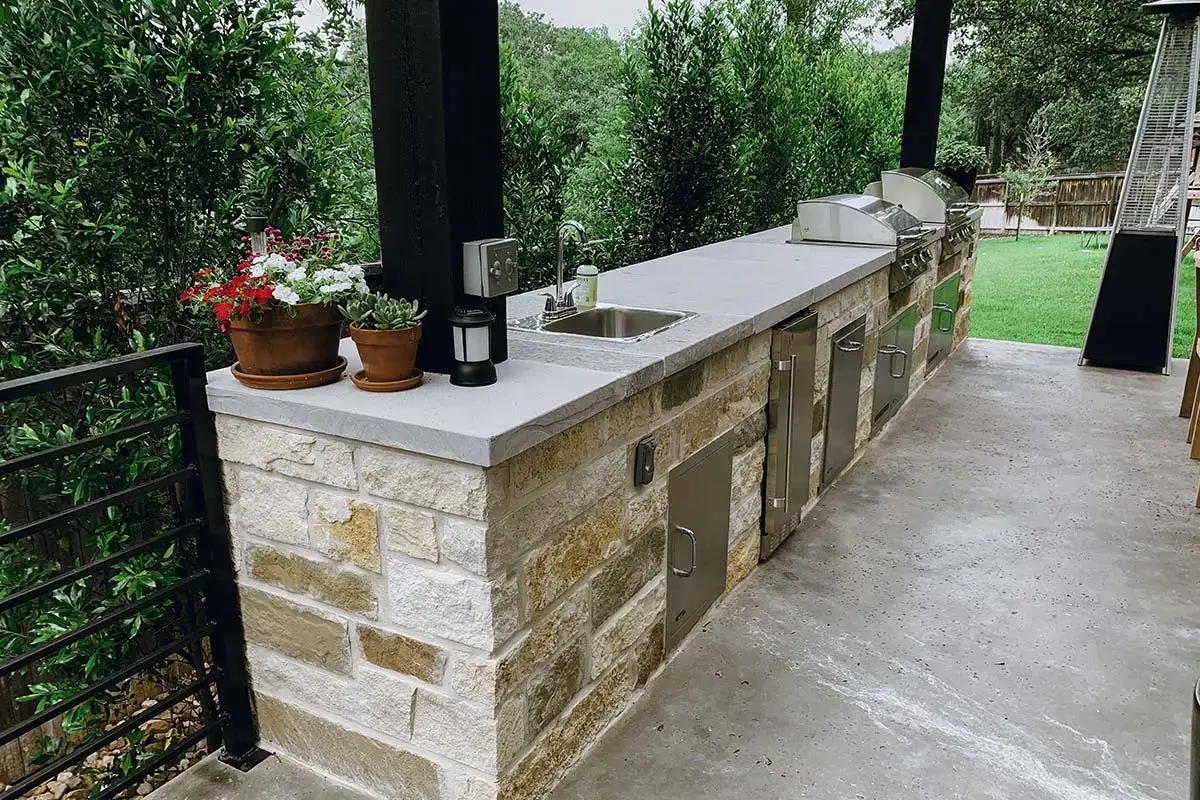 Outdoor kitchens with a stone countertop, built-in grill, sink, and stainless steel cabinets, surrounded by greenery