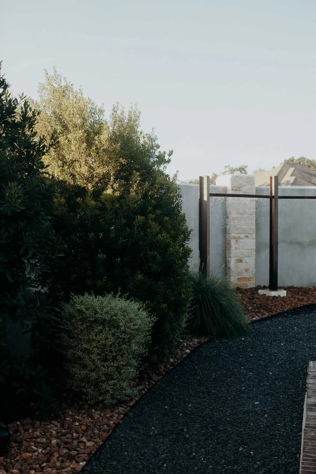 Gravel path lined with lush green softscapes, including bushes and trees, leading to a minimalist water feature