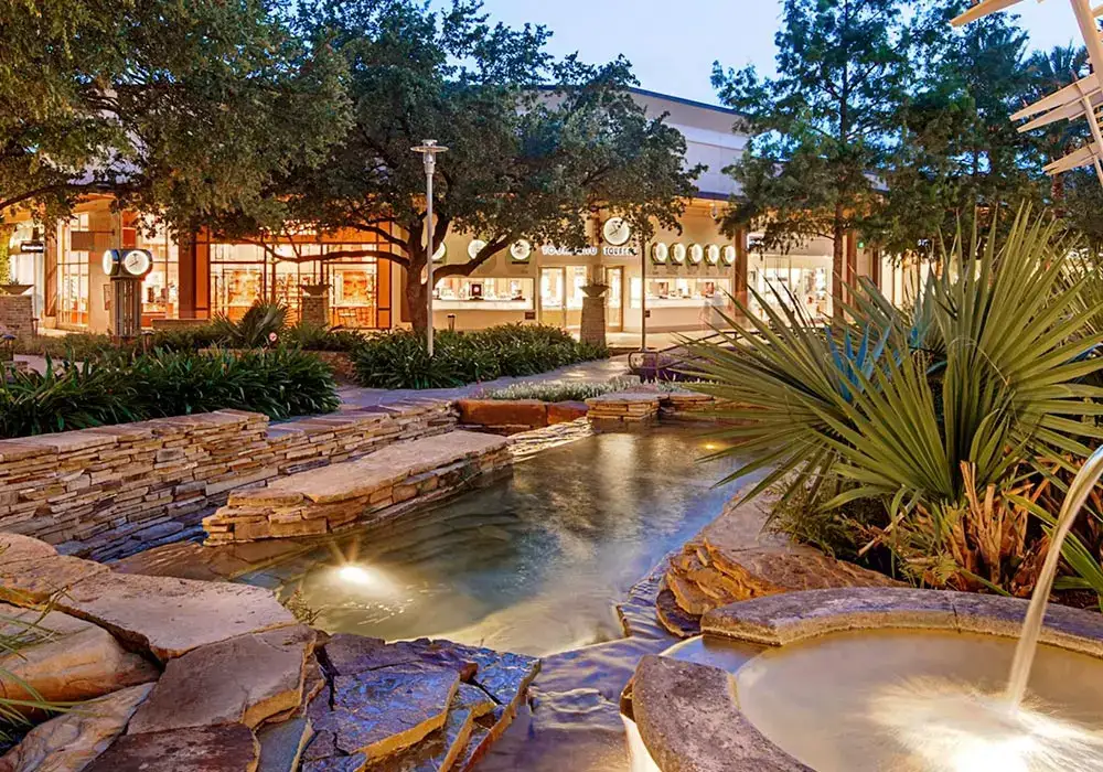 Commercial lanscape with stacked stone walls, illuminated water features, and lush greenery surrounding a retail area