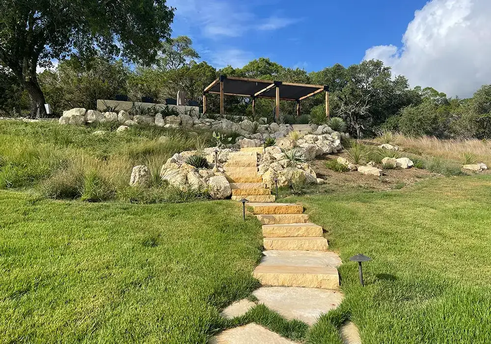 Stone steps leading up a grassy hill to a shaded pergola, surrounded by natural boulders and integrated hardscapes