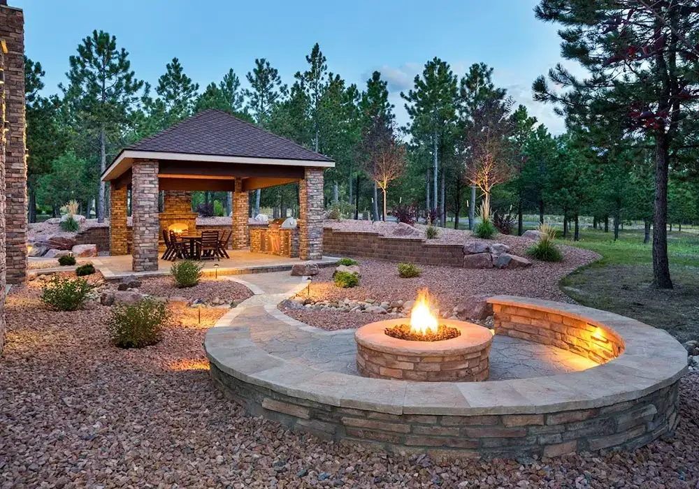 Outdoor kitchens with a stone pavilion, built-in grill, and fire pit, surrounded by a gravel landscape and pine trees