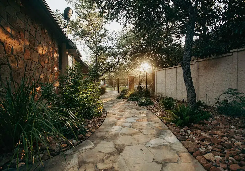 Stone pathway in a residential lanscape, bordered by lush plants and trees, leading to a gate with soft evening light