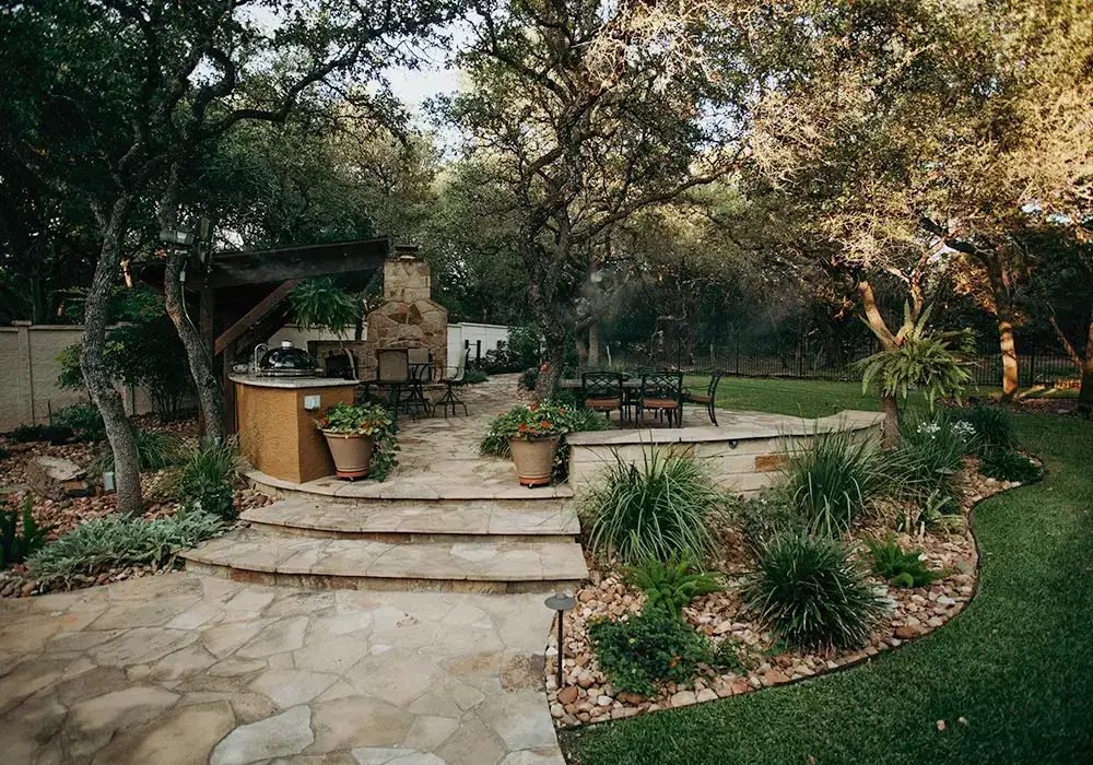 Outdoor kitchens with a built-in grill, stone countertops, and fireplace, surrounded by trees and landscaped greenery