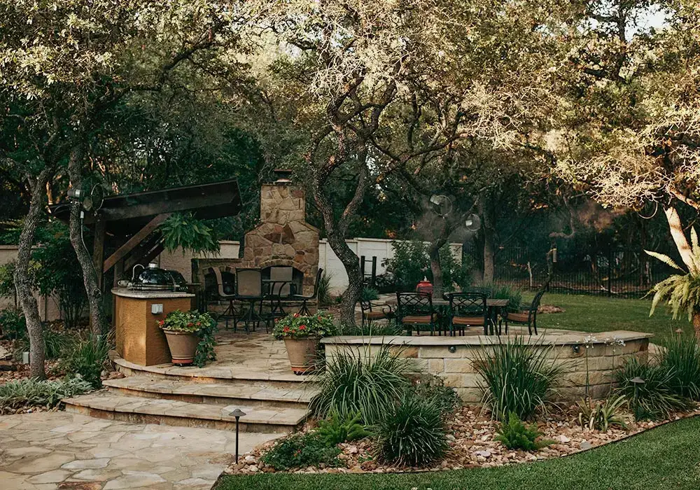 Outdoor kitchens with a built-in grill, stone fireplace, and seating area, surrounded by trees and lush landscaping