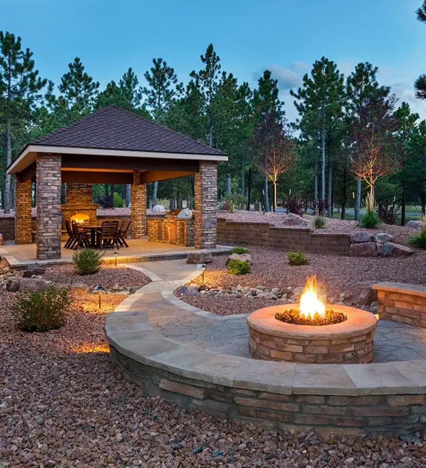 Outdoor pavilion with a fire pit, stone hardscapes, and a cozy seating area surrounded by trees and lighting